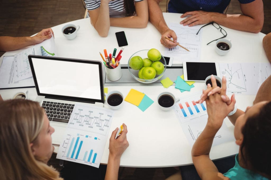 A group of people are seated around a table with laptops, charts, and documents. The table has a centerpiece with green apples, cups of coffee, and office supplies like pencils and sticky notes. Some figures and graphs are visible on papers and electronic devices.