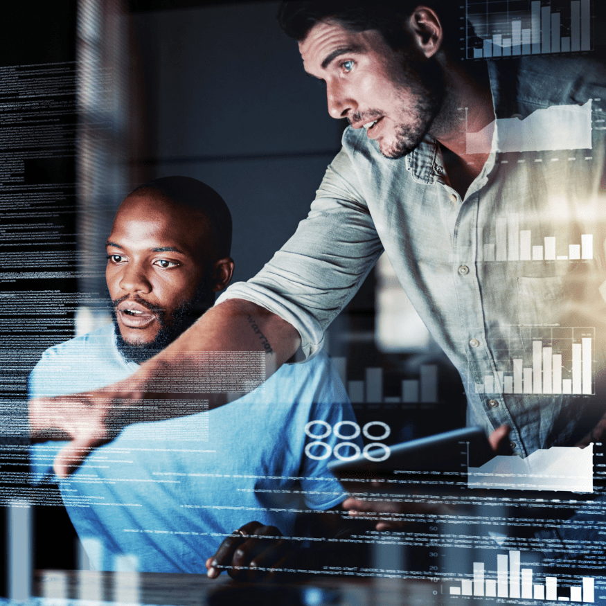 Two men are working together in an office environment, surrounded by digital charts and graphs projected on glass. One man is pointing at the data, while the other looks intently at the information, suggesting a collaborative effort in analyzing or discussing the data.