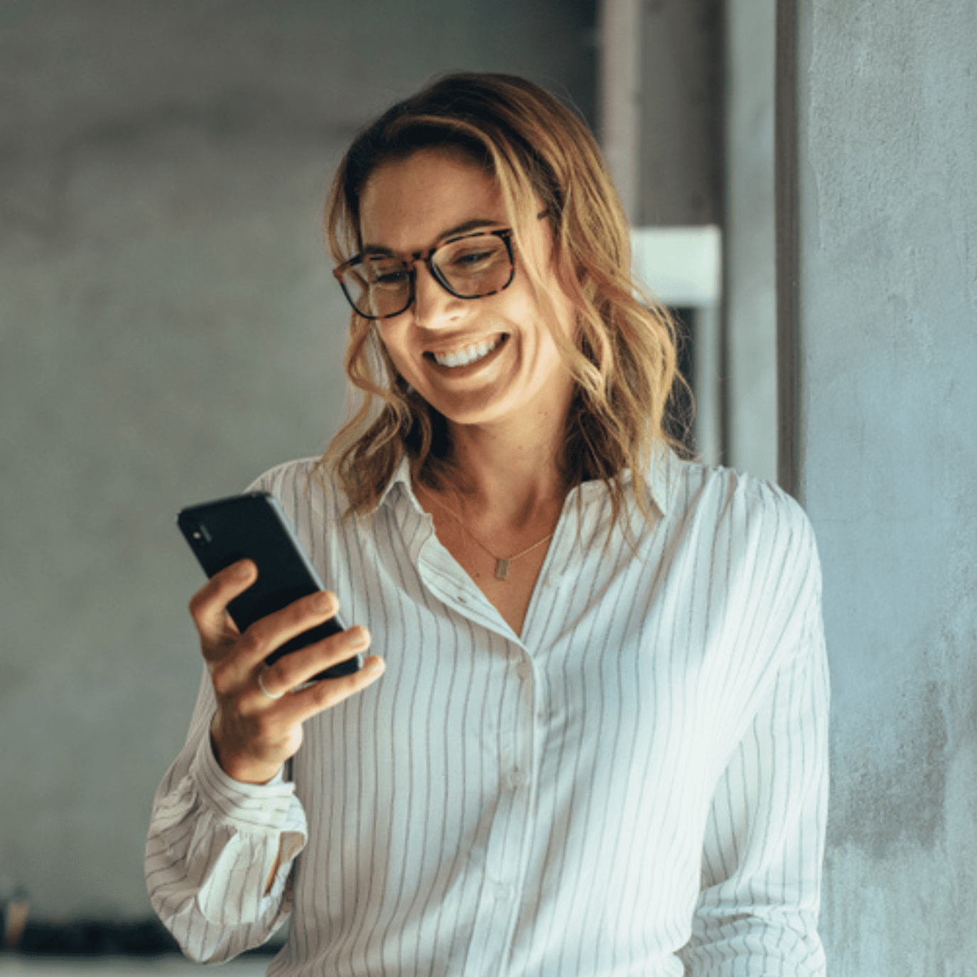 A woman with glasses and a striped shirt stands smiling while looking at her smartphone. She is leaning against a wall, appearing to be in a bright indoor space.