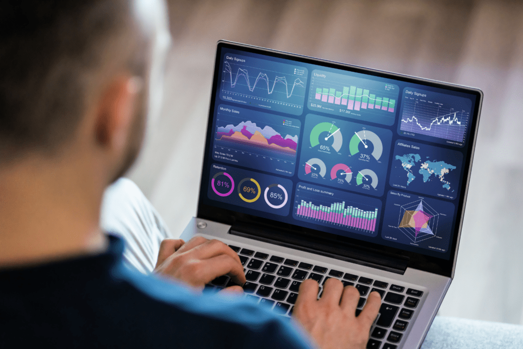 A person using a laptop displaying various colorful data visualization charts and graphs related to business analytics, including line graphs, pie charts, bar graphs, and world maps with data points. The person is typing on the keyboard.