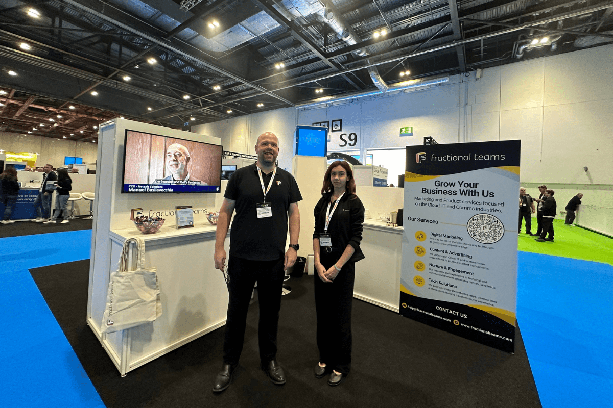 Two individuals stand at a trade show booth for "Fractional Teams,". The booth features a TV screen, a vertical banner detailing services like digital marketing, and a small table with promotional materials. The venue boasts a modern, industrial appearance.