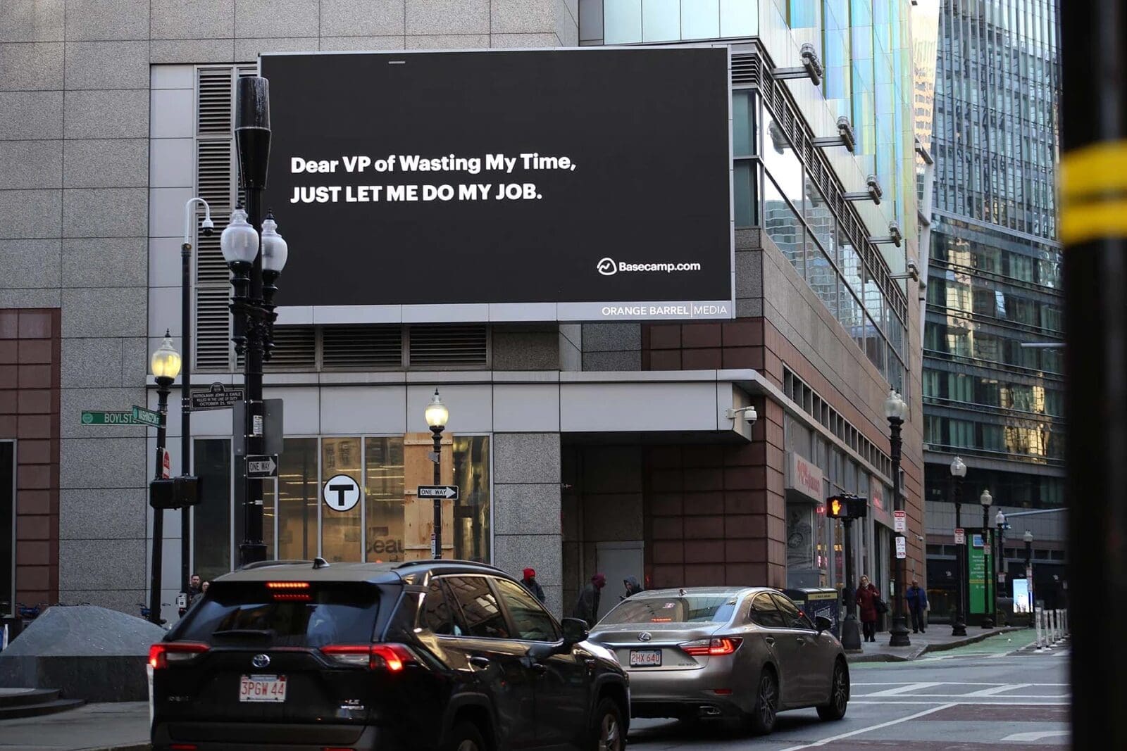 A billboard on a city street reads, "Dear VP of Wasting My Time, JUST LET ME DO MY JOB. Basecamp.com." Perfectly capturing the essence of clever content marketing, two cars sit in the foreground amidst a daytime scene with modern buildings and a streetlamp visible.